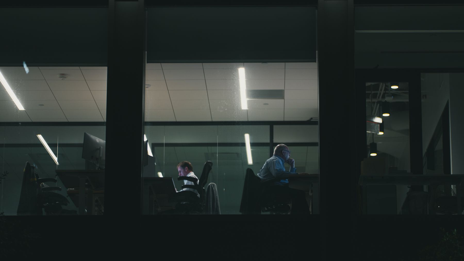 Two professionals working at separate desks inside a glass-walled office at night.